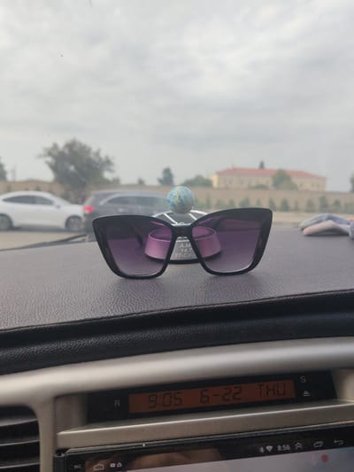 The image shows a pair of black cat-eye sunglasses with purple lenses resting on a dark, textured dashboard inside a car. A small, blue and white marbled globe ornament is positioned behind the sunglasses, and to the right, a grey and white patterned fabric item is visible. In the background, through the car's windshield, a street scene is blurred. Several cars are on the road, including a white sedan and a dark SUV. Beyond the road, a building with a red roof and light-colored walls is visible, surrounded by trees and a light-colored wall. The sky is overcast. On the dashboard, a digital display shows the time as "9:05" and the date as "6-22 THU". The car is likely in Baku City, Azerbaijan, given the presence of the car's dashboard display showing the time and date in a format common for the region.