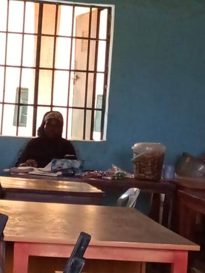 A woman wearing a black garment and a patterned headscarf sits behind a wooden table in what appears to be a classroom or office setting in Kakuri Gwari, Nigeria. Sunlight streams through a barred window, illuminating the room. On the table in front of the woman are several papers, a tissue box, and a dark object, possibly a phone. To her right, on a separate table, a clear plastic bucket filled with food items is visible, along with other objects that are difficult to discern. The room has a blue wall and multiple wooden tables are present.