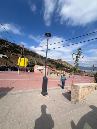 The image depicts a daytime scene in Bin El Ouidane, Morocco. A paved walkway transitions from red to gray tile. A black lamppost stands prominently on the gray tiled area. A mountain rises in the background, with buildings and trees visible along its slopes. A building with signage and posters stands adjacent to the red paved area. A yellow sign is propped up by wooden supports next to parked cars. Utility lines run across the sky. A person in a blue jacket walks in the middle ground. The sky is blue with scattered clouds. A shadow cast by a person taking the photo is visible in the lower right corner.