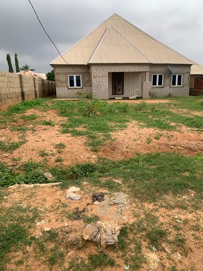 This image depicts a residential property in Kakau, Nigeria, under an overcast sky. A light-colored, modern house with a metal roof dominates the center of the frame. The house has a porch with a wooden door and two windows with dark panes on the front. There are two windows on the right side of the house, also with dark panes. Red graffiti is visible on the wall of the house. To the left of the house is a brick wall, and beyond that, two tall, thin trees are visible. The yard in front of the house is a mixture of sandy soil and patchy green grass, with some areas appearing to be undergoing construction or landscaping. Scattered debris, including plastic bags and what appears to be discarded construction material, is present in the foreground of the yard. To the right of the main house, another structure with a similar metal roof and a dark wooden door is partially visible. A blue satellite dish is mounted on the wall of the main house. The overall impression is of a developing residential area.