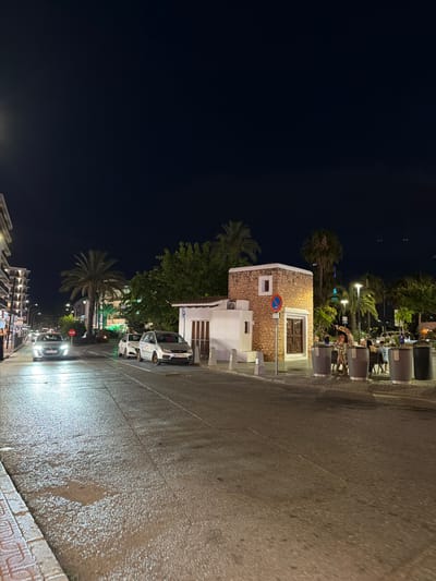 The image captures a street scene at night in Sant Antoni De Portmany, Spain, likely after a rain shower given the wet pavement reflecting the streetlights and headlights. The perspective is from the street looking towards a building with a stone facade and a white section.

The building has a distinctive arched window with decorative metalwork. A small rectangular window is also visible on the stone part of the building, and an air conditioning unit is mounted on the white section. A circular blue and red sign with a white diagonal bar is affixed to the stone wall.

Several cars are parked and driving on the wet street. Some are illuminated by headlights, creating bright reflections on the asphalt. In the background, palm trees and other vegetation are visible, illuminated by streetlights. Further back, there appears to be a row of buildings with lights on, suggesting shops or residences.

People are sparsely visible, primarily in the background, seated at tables outside what seems to be a restaurant or cafe. They are casually dressed, and their presence contributes to a lively, albeit subdued, nighttime atmosphere. The overall mood is tranquil and somewhat damp, typical of a coastal town after dark and a recent rainfall. The scene suggests a typical evening in a popular tourist area. No discernible text is clearly readable in the image.