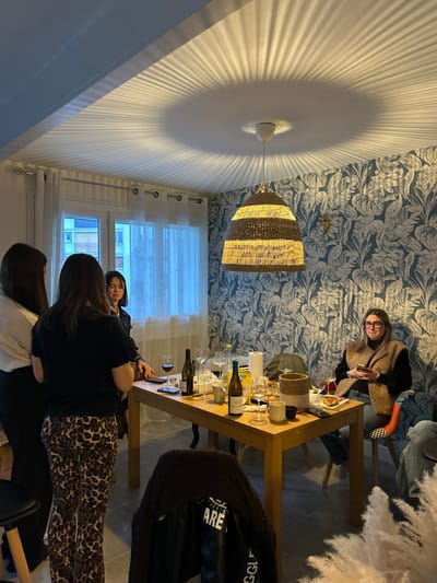 A casually set dining table displays wine bottles, glasses, and scattered items, suggesting a recent gathering. Three people are near the table, one in leopard-print pants and a black top, another in black, and a third in white. A woman sits at the table's far end, wearing glasses and a tan sweater. A distinctive lamp casts radiating shadows on the ceiling. Wallpaper with a blue leafy pattern adorns the wall, and natural light filters through a window.