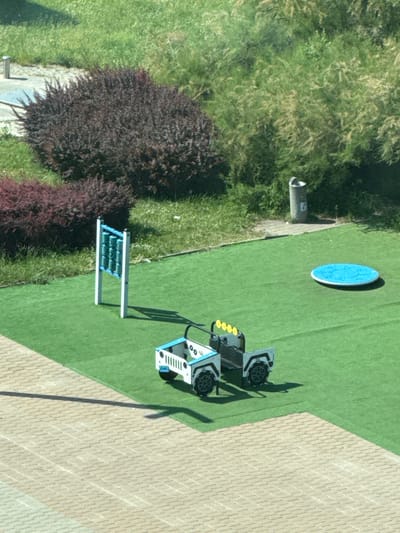 The image depicts an outdoor children's play area with green artificial turf. In the foreground, a light-colored toy jeep with blue accents and four yellow headlights is parked on the turf. To the left of the jeep, a blue and white musical xylophone-like structure stands vertically. Further back, a dark red bush is prominent, with a lighter red bush to its left. Behind the bushes, a trash can is visible. In the background, lush green grass and trees dominate the scene. The play area appears to be part of a larger urban environment, possibly a park or residential complex. The lighting suggests it is daytime, with shadows cast by the objects. The image does not show any people. The location is identified as a playground in Gdynia, Poland.