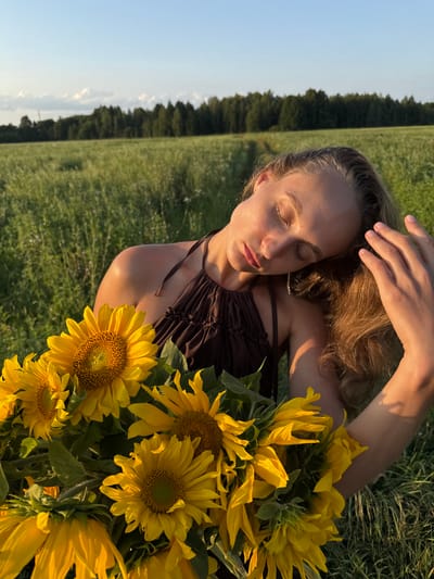 A young woman with blonde hair is in a field of green grass holding a bouquet of sunflowers. She is wearing a brown halter top and her eyes are closed as she tilts her head back. The sun is setting, casting a warm glow on the scene. In the background, there is a line of trees and a clear blue sky. The image appears to have been taken in Aksaitovo, Russia.