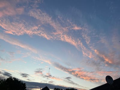 This is an outdoor, eye-level, wide-angle photograph taken at sunset in the city of Sredny, Russia. The sky dominates the frame, filled with a gradient of light blue to a deeper blue towards the top. Wispy, elongated clouds are scattered across the sky, tinged with soft pink and orange hues from the setting sun. In the lower part of the image, the silhouettes of several rooftops are visible, some with dark trees in front of them. A satellite dish is mounted on one of the rooftops on the right side of the frame, and an antenna or tall pole is visible on the left. The lighting suggests it is either dawn or dusk.