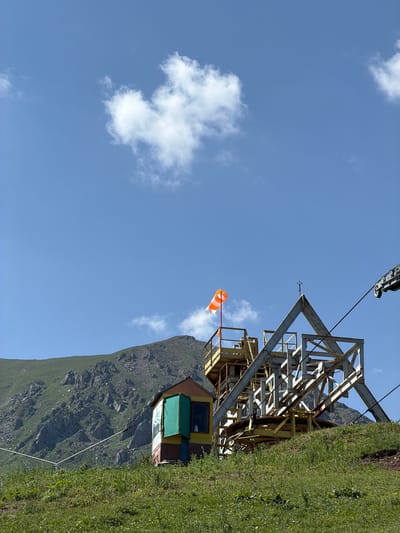 In Kashka Suu, Kyrgyzstan, an orange and white windsock flutters atop a metal structure on a grassy hillside. The structure, likely part of a ski lift or gondola system, features yellow and white beams and platforms. A small, colorful building with green shutters stands to the left of the main structure. Beyond the structures, a rugged, green mountain range dominates the background under a clear blue sky dotted with white clouds. A cable stretches across the frame, suggesting an operational lift system. The ground is covered in lush green grass with scattered yellow wildflowers.