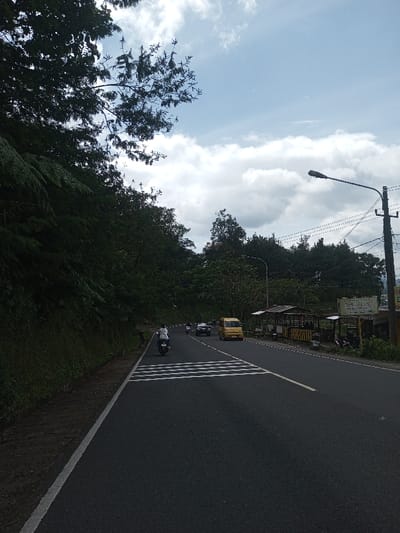A wide-angle, slightly elevated perspective captures a two-lane asphalt road curving gently to the right, flanked by dense vegetation and roadside structures under a partly cloudy sky, in the city of Gubbisamamidi, India.
To the left, a steep hillside is covered in dense, dark green foliage, including ferns and trees, extending upwards out of frame. The asphalt road features a solid white line marking its left edge and a solid yellow line separating the two visible lanes. In the foreground, a pedestrian crosswalk is marked by four parallel white stripes across the left lane.
A single individual, wearing a white shirt and dark pants, operates a white scooter on the left lane, moving away from the viewer towards the crosswalk. Further down the road in the right lane, a yellow van is observed, followed by additional cars and motorcycles receding into the distance.
On the right side of the road, a roadside stall with an open-air design is present, featuring a corrugated metal roof supported by wooden posts. A yellow sign, displaying text that includes "SANGKAR KUSUMA," "TEH JAWA," and "KOPI NAGA," is affixed to the stall. Several motorcycles are parked near this structure. A utility pole, equipped with a streetlamp and multiple power lines, stands prominently on the right. The background on the right consists of more lush trees and distant vegetation. The sky is partly cloudy with substantial white clouds and interspersed patches of blue.