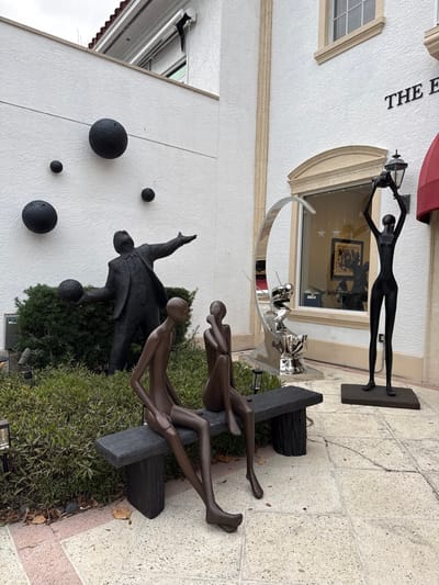 The image displays an exterior scene in Naples, United States of America, featuring a building facade with multiple sculptures. The structure consists of white stucco walls with light-colored architectural trim around windows and a vertical fluted pilaster. A portion of a red tile roof is visible at the top left, and a red awning extends from the right side. A dark sign with the partially visible text "THE E" is mounted on the wall above an arched window.
The foreground contains a dark-colored bench supporting two stylized, slender, dark reddish-brown figures. The figure on the left, male, is seated with legs outstretched and hands between the knees. The figure on the right, female, is seated with legs crossed at the ankles, a hand near her chin. These figures are situated within a planter bed densely populated with green bushes, which also conceals the lower portion of a dark-colored male figure positioned behind them. This male figure, dressed in a suit, stands with his head tilted upward, one arm extended holding a black sphere, and the other arm raised.
Attached to the white wall behind these sculptures are four black spherical objects of varying sizes. A black security camera is mounted high on the wall to the upper left. To the right of the sitting figures, a large, reflective silver-colored abstract sculpture, resembling a crescent moon, stands prominently before the arched window. Inside this window, white star decorations and a dark-framed artwork are visible. Further right, a tall, slender, black figure stands on a dark square base, holding a black lantern with both hands above its head. The ground is paved with light-colored square tiles, with a few dry leaves scattered near the planter. The sky above is overcast.