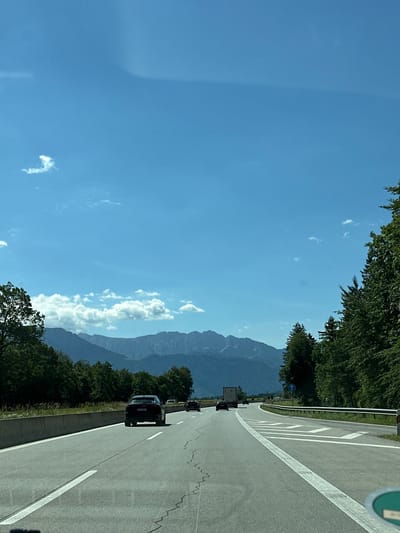 The image depicts a sunny day with a clear blue sky and scattered white clouds. A highway stretches through a landscape with lush green trees on either side and a majestic mountain range in the background. Several cars and a truck are visible on the road, indicating traffic. The presence of a highway, mountains, and dense vegetation suggests a scenic route, possibly in a rural or semi-rural area. Given the context of Flintsbach am Inn, Germany, this scene likely captures a view of the Bavarian Alps.