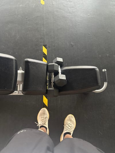 The image provides a top-down view within a fitness facility in Santa Catarina, Mexico. The floor is dark gray, marked by a prominent yellow and black striped line running vertically through the center. A black, adjustable weight bench with a silver-colored frame is positioned horizontally across this floor marking. Two hexagonal black dumbbells rest on the padded surface of the bench, centered between its two main sections. In the foreground, the lower torso, legs, and feet of a person are visible. The individual is wearing dark athletic shorts, light-colored athletic shoes with textured soles, and dark socks, with their feet positioned on either side of the yellow and black floor line.