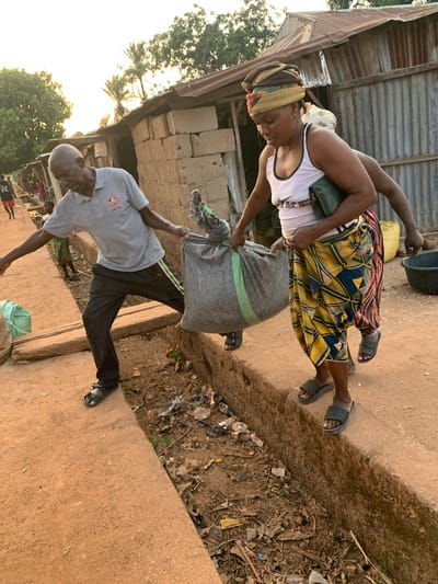 In Agban, Nigeria, two individuals interact outdoors. A man in a light grey t-shirt and dark pants extends his arm towards a large grey bag held by a woman. The woman, wearing a white tank top, a patterned yellow and blue skirt, and a head wrap, appears to be handing over the bag. The setting includes a building with a corrugated metal roof and a partially visible brick wall. The ground is a mix of dirt, debris, and a concrete path. Nearby, there are trees.