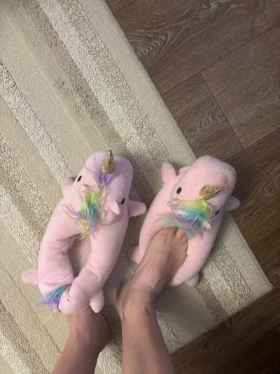 The image shows two pink unicorn slippers, one worn by a person's left foot and the other by their right foot, on a striped rug. The slippers have gold horns and colorful manes. The person's feet and ankles are visible. The background consists of a wooden floor. The lighting suggests it is daytime. The location is Voronezh, Russia.