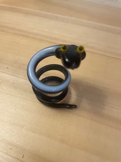 A glass figurine of a coiled black and grey snake with yellow eyes sits on a light-colored wooden surface. The snake is coiled into a spiral shape, with its head at the top and its tail extending downwards and to the right. The surface appears to be a table or countertop with wood grain visible. There is no indication of the city of Taganrog, Russia.