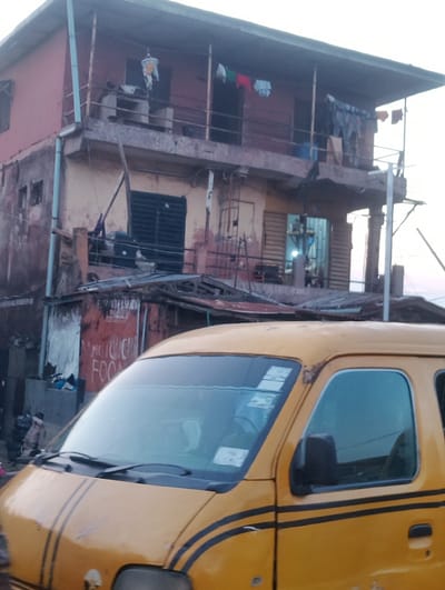 The image captures a scene on a street in Akowonjo, Nigeria. In the foreground, the front portion of a yellow commercial vehicle, likely a minibus, is visible. The vehicle has black stripes along its side and appears to be parked or moving slowly. The windshield and side windows of the vehicle are tinted. Behind the vehicle, a multi-story building with a weathered facade is depicted. The building has balconies with railings, and laundry is hanging out to dry on clotheslines. One balcony has a hanging light fixture with clothes on it. On the ground floor of the building, a small, makeshift structure with a red painted exterior is visible, with text that appears to read "MUCH FOOD". The overall environment suggests a densely populated urban area. The lighting in the image indicates it may be either early morning or late afternoon.
