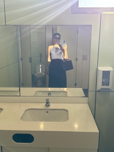 In a restroom, a person is taking a mirror selfie. They are wearing a dark dress and sunglasses and are holding a smartphone and a laptop. The restroom has white tiled walls and a white toilet. There is a large mirror covering one wall, reflecting the scene. A window with sunlight streaming through is visible in the upper left corner. The overall setting appears to be a public restroom, possibly in an airport or a similar facility, given the functional design and the presence of a laptop. The lighting suggests it is daytime.