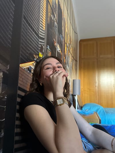 A young woman lies on a floor with a photorealistic city grid design, covering her mouth with her hand. She wears a black top, a gold watch, and a gold bracelet. A large wooden cabinet is visible in the background along with a blue blanket and exercise equipment. The location in Mirandela, Portugal is not identifiable from the image.