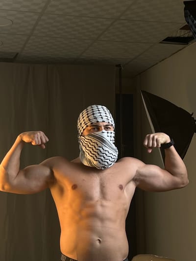 A muscular, shirtless man poses in what appears to be a studio setting. He wears a keffiyeh over his head and face, obscuring all but his eyes. His right arm is raised, flexing his bicep, and a watch is visible on his wrist. His left arm is also flexed downwards. A softbox studio light is positioned behind his right arm. The background features a wall with a tiled pattern.