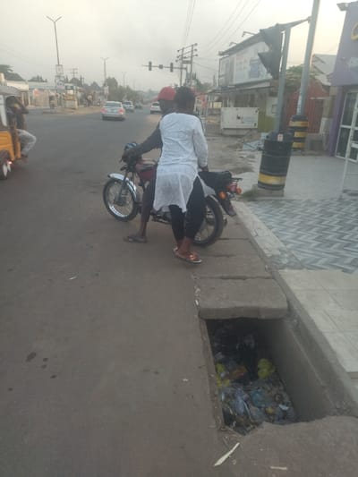 The image shows two people standing next to a motorcycle on the side of a paved road in New Karu, Nigeria. The motorcycle is facing the road, and the two people are standing next to it, with one person appearing to be getting off. The person getting off is wearing a white long-sleeved shirt and appears to be a woman. The other person is wearing a red shirt, and appears to be a man. Both individuals have their backs to the camera. The motorcycle is parked on the sidewalk, which is made of concrete. The sidewalk is next to the road, which is paved with asphalt. There is a large drain on the sidewalk that is filled with trash. The road is wide and straight, and there are buildings on either side of it. The sky is overcast, and the ground is dry. There are several power lines running above the street, as well as one traffic light that is not active. The road is empty of vehicles, except for the motorcycle, a yellow-green vehicle, and a white sedan in the background. The time of day is either early morning or late afternoon, as the sky is somewhat hazy and there are long shadows. There is no vegetation in the scene, and the ground is dry and dusty. The overall scene is a typical urban street in a developing country. There is a sign in the background which advertises a business that sells phones, accessories, and airtime. A trash can on the sidewalk is also visible in the background. The picture appears to be taken from a position on the sidewalk, looking down toward the street.