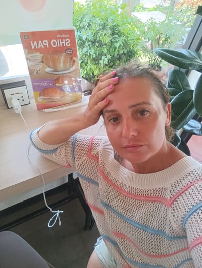 A woman is sitting at a table indoors, possibly at a restaurant or cafe. She has her hand on her forehead and is looking at the camera. She is wearing a white sweater with pink and blue stripes. On the table next to her is an advertisement for Shio Pan, a type of bread, in Thai. There is also a power outlet with a charger plugged in. Outside the window, there are green plants and trees. The location appears to be Plai Phraya, Thailand, based on the Thai writing on the advertisement.