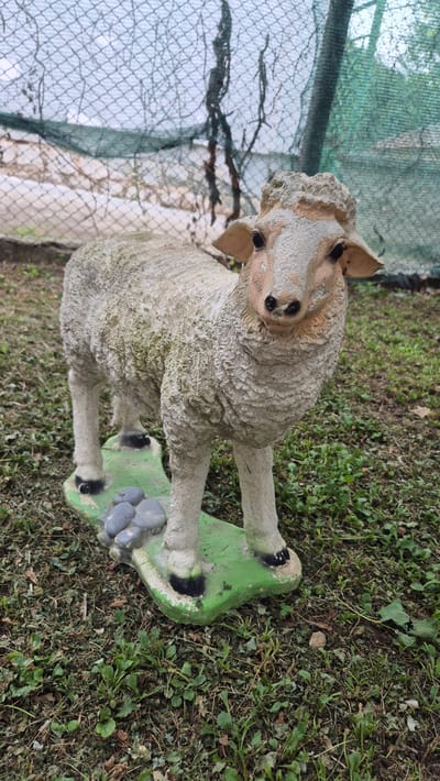 A concrete statue of a sheep stands on a green, irregular-shaped base with several gray stones on it. The sheep statue is white with some dirt and moss on it. It has black eyes and black hooves. The base is placed on the ground, which is covered with dry leaves and green grass. In the background, there is a green mesh fence. The scene is outdoors. The image was taken in the city of Doirentsi, Bulgaria.