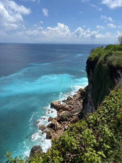 The image depicts a coastal scene in Uluwatu, Indonesia. A rocky cliff covered in dense green vegetation extends over the turquoise ocean. Waves crash against the rocks below. The sea transitions from a lighter, more vibrant blue near the shore to a darker, deeper blue further out. The sky, visible to the left, features a blue expanse with scattered white clouds.