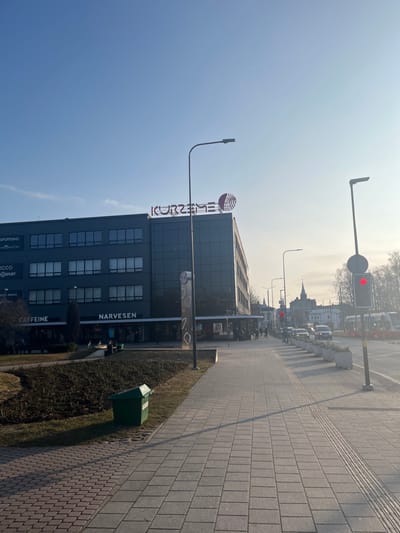 This image depicts an urban street scene in Liepaja, Latvia, specifically near the Kurzeme shopping center at Lielā iela 13. The scene is observed on a clear, sunny day, casting long shadows.
In the foreground, a light grey paved sidewalk with tactile paving strips extends from the lower left into the distance. To the left of the sidewalk, a patch of sparse grass and low, dormant bushes contains a green, rectangular public trash bin.
The central subject is a multi-story modern building with a dark grey facade and numerous glass windows, identified by a large red "KURZEME" sign and stylized logo on its rooftop. Ground-level signage on this building includes "NARVESEN," "CAFFEINE," "SPORTLAND," "H&M HOME," and "ECCO." Several pedestrians are visible walking on the sidewalk in front of the building and near its entrance. A tall, dark grey streetlamp stands in front of the building, to the left of the main path.
To the right, a parallel street is visible with ongoing vehicle traffic; several cars are present, some with illuminated headlights. This street is separated from the sidewalk by a row of rectangular concrete planters containing low green vegetation. Further along the sidewalk on the right, another streetlamp and a prominent traffic light displaying a red signal are present.
In the background, under a clear blue sky, a church spire or similar architectural structure is visible in the distance, along with other urban buildings and what appear to be public transport vehicles.