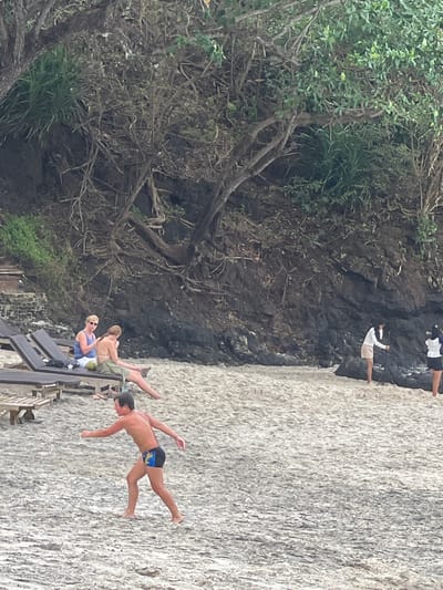 The image depicts a beach scene in Manggis, Indonesia. The foreground features a sandy beach with a few lounge chairs. A young person in black and blue swim trunks is captured mid-air, appearing to jump or dive. Further back, a rocky cliff face is visible, with some vegetation growing on it. Two people are standing on the cliff, one in a white shirt and beige shorts, and another in a black and white top and black shorts. Another person is visible higher up on the cliff, their body obscured by the foliage. In the lower left of the frame, a person wearing sunglasses and a blue top is sitting on a lounge chair, and another person in a bikini is partially visible next to them. The scene appears to be a daytime activity, with bright natural light illuminating the beach and cliffs. The overall atmosphere suggests a relaxed, recreational environment at a tropical beach. There is no visible text in the image.