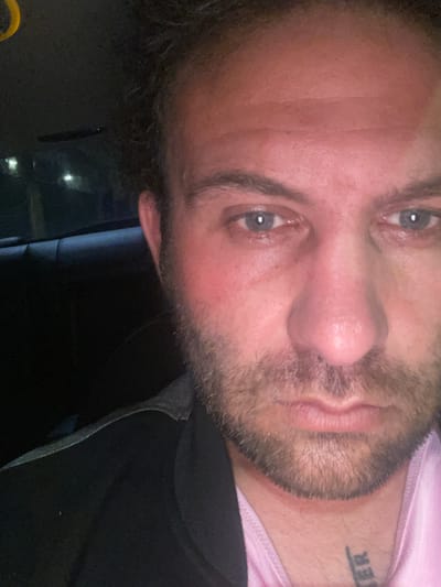 A close-up selfie of a man with a short beard and light eyes. He is wearing a pink shirt under a black jacket. A tattoo is visible on his neck. The location is unknown, and there is no indication it is in Armagh, United Kingdom.