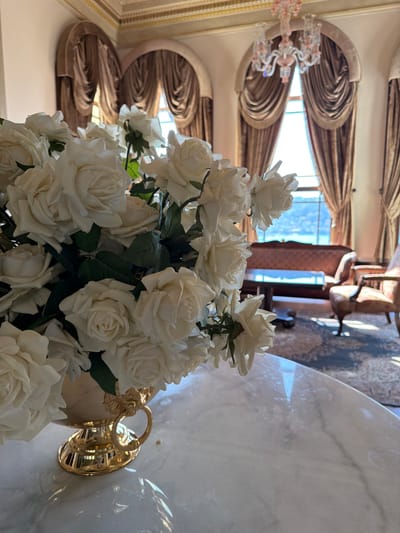A bouquet of white roses in a gold vase sits on a marble table in a sunlit room with large arched windows draped with heavy brown curtains. In the background, a view of the Bosphorus strait in Üsküdar, Turkey, is visible through the windows, with a brown sofa and armchair placed on an oriental rug. A chandelier hangs from the ornately decorated ceiling.