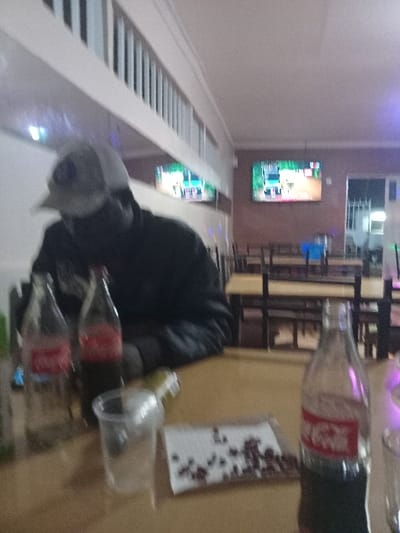 The image displays an indoor entertainment setting, possibly a bar or club, located in Chevaywa ward, Kenya. The scene suggests an evening or nighttime atmosphere, illuminated by artificial lighting, which includes a purple glow in some areas.
In the foreground, a person is visible, dressed in a dark jacket or hoodie and a light-colored cap, looking downwards towards a table or counter. This light brown surface holds several glass bottles; two are clearly identifiable as Coca-Cola bottles due to their shape and red labels, though the text is blurry. Another clear plastic bottle or glass also rests on the table, along with a white paper or napkin featuring a dark, speckled pattern.
A prominent feature in the midground is a long, light-colored structure with parallel lines, resembling a bowling lane or a similar indoor game facility. This surface reflects light and images from the surroundings. Mounted on the far wall are two brightly lit, rectangular electronic displays showcasing vibrant, colorful graphics, likely from video games or dynamic visual content. Blurred dark furniture, possibly stools or chairs, can be seen in the background. The overall ambiance suggests a casual social gathering or leisure activity.