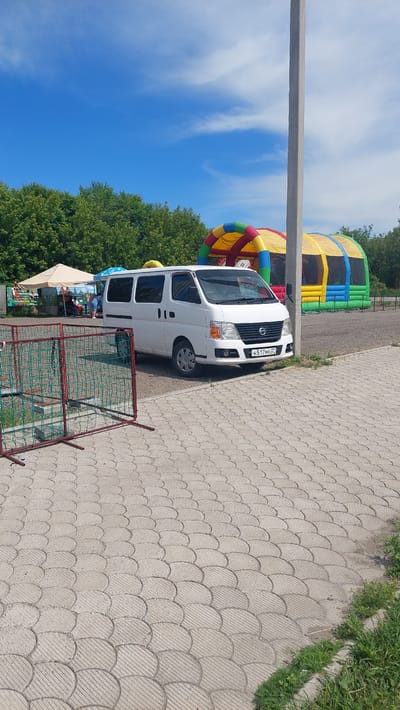 The image depicts an outdoor scene in Komsomolsk-on-Amur, Russia, on a sunny day with a blue sky and scattered clouds. In the foreground, a light-colored, multi-paneled paved area leads to a white van. The van, a Nissan NV200 or similar model, is parked on the paved area and faces slightly towards the right. Its license plate is partially visible, displaying the characters "H517MA 87". To the left of the van, a red-fenced play area with green netting is visible.
In the background, there are several elements suggesting a recreational or event setting. A large, inflatable structure, partially yellow, red, and green, stands prominently behind the van. To the left of this structure, a light-colored tent or canopy is set up, with people milling about underneath and around it. Further left, a line of lush green trees forms the background. A tall, grey concrete utility pole stands to the right of the van, extending upwards out of the frame. The overall impression is a scene from a park, fairground, or event space on a bright, clear day.
