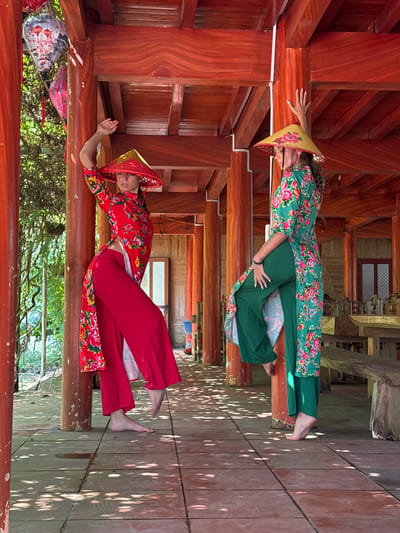 Two women are captured in a posed photograph, lying horizontally on robust, reddish-brown wooden beams within what appears to be a traditional Vietnamese wooden structure, possibly a house or pavilion in Thông Nguyên Commune, Vietnam.

The woman positioned higher in the frame wears a vibrant green áo dài, a traditional Vietnamese long tunic, adorned with a floral pattern of red and white blossoms, paired with matching green trousers. Her head is covered by a woven conical hat (nón lá) with a yellow trim. She lies on her back, with her left arm bent, hand resting gently on her stomach, and her right arm extended slightly.

Below her, the second woman is dressed in a striking red floral áo dài with red trousers and a red nón lá. She also lies on her back, her left hand resting on the beam near her head, while her right arm is bent at the elbow, hand near her waist. Both women are barefoot and appear serene, looking towards the camera with calm expressions.

The setting is characterized by highly polished wooden beams and planks, forming the structure around and beneath them. Below the women, a wooden floor is visible, marked by dappled sunlight and shadows, suggesting a bright, sunny day. Further down and to the left, decorative lanterns hang, and green foliage or trees can be seen, indicating an outdoor or semi-outdoor location. The intricate woodwork and traditional attire are strong cultural cues of Vietnam. No text is visible in the image.