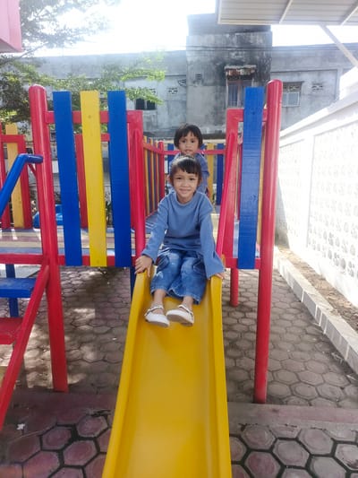 The image displays an outdoor scene in Pantegajah, Indonesia, featuring two young children on a multi-colored playground structure under bright daylight. In the foreground, a child, likely of East Asian descent, is seated on a yellow slide, facing the camera with a smile. This child is wearing a light blue long-sleeved shirt, blue jeans, and white flat shoes with straps. Behind and slightly above, a second child, similarly attired in a light blue long-sleeved shirt and blue jeans, is partially visible from the chest up, positioned further back on the playground structure and looking towards the camera.

The playground equipment consists of a yellow slide, red structural supports, blue steps, and vertical barriers made of alternating blue and yellow planks. The ground is paved with hexagonal concrete blocks, exhibiting variations in color from grey to reddish-brown, with some darker, potentially damp or shaded areas. In the background, a weathered, multi-story grey concrete building with visible windows and signs of discoloration or rust is present, partially obscured by sparse green foliage. To the right of the playground, a white wall with a repeating circular pattern in relief extends, alongside a segment of a white metal awning structure.