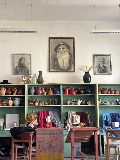 The image captures an interior wall in Troyan, Bulgaria. On the wall, three framed portraits hang vertically, two of older women and one of an older man with a beard. A dark-colored vase containing dry foliage is positioned between the upper two portraits. A light fixture is mounted vertically on the left side of the wall. To the right of the portraits, a tall, narrow, multi-tiered shelving unit, painted green, displays an eclectic assortment of objects: pottery, teapots, kitchenware, textiles, and a few glass bottles. A series of chairs, some stacked, are partially visible to the right of the shelving unit.