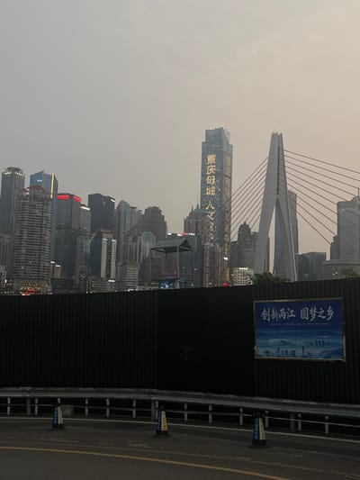 The image captures a wide shot of a city skyline at dusk, with a focus on a modern bridge and a cluster of skyscrapers. The sky is overcast and hazy, casting a muted light over the scene. In the foreground, a dark, ribbed fence runs horizontally across the image, partially obscuring the view of the city. A road with yellow and white lane markings is visible in the lower portion of the frame, with two traffic cones placed near the fence.
Dominating the center-right is a distinctive cable-stayed bridge with a single, large white pylon. The cables extend outwards, connecting to the bridge deck. To the left of the bridge, a tall, slender skyscraper is illuminated with vertical lines of yellow text. The text appears to be Chinese characters. Behind this prominent building are numerous other skyscrapers of varying heights and architectural styles, some with illuminated windows and signs. One building towards the far left has "ADBC" visible on its upper section.
On the fence in the lower right quadrant, a large blue and gold framed poster is affixed. The poster displays an artistic rendering of a cityscape with buildings and a body of water, along with Chinese text at the top. The location is identified as 江北城街道 (Jiangbei Cheng Subdistrict), China. The overall impression is one of urban development and modern infrastructure in a coastal or riverside setting, seen through a hazy atmosphere.