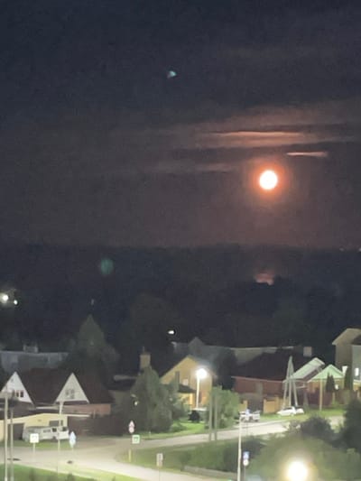 The image depicts a nighttime aerial view of a residential area in Laishevo, Russia, under a full moon. The moon, a vibrant orange sphere, is partially obscured by thin clouds, casting a soft glow over the landscape. Below, a cluster of houses with illuminated windows and dark rooftops is visible, nestled amongst trees and a winding road. A street light illuminates a section of the road, and several parked vehicles are scattered throughout the scene. The overall atmosphere is serene and quiet, characteristic of a peaceful evening in a suburban setting. In the sky, a faint, teal-colored object is visible to the left of the moon.