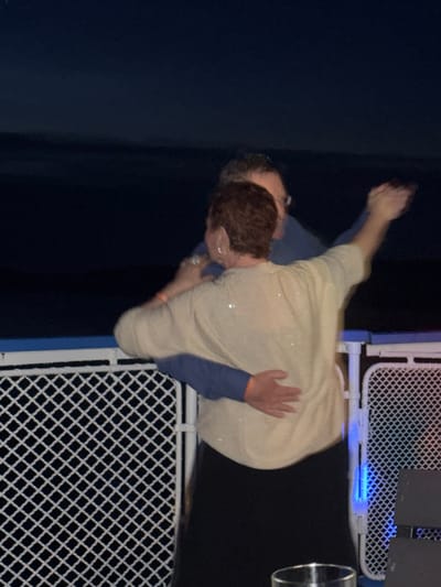 Two individuals are embracing and dancing on what appears to be a boat deck at night. The woman, with short reddish-brown hair and wearing a sparkly cream-colored top and a black skirt, has her arms around the man's neck. The man, wearing glasses and a blue shirt, has his arms around the woman's waist and is holding her hand. A white chain-link fence with blue lights is visible in the foreground, and a dark sky with a faint horizon line is in the background. The location of Dulesovo, Russia is not determinable from the provided image.