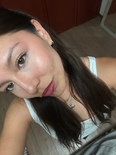 A close-up shot of a young woman's face, angled toward the camera. She has brown eyes, brown hair, fair skin, and is wearing lipstick. A gold earring is visible in her left ear, and a necklace with a heart-shaped pendant is around her neck. She is wearing a light-colored top. The background shows a floor and possibly a bed. There is no information in the image to confirm that it was taken in Heroica Puebla de Zaragoza, Mexico.