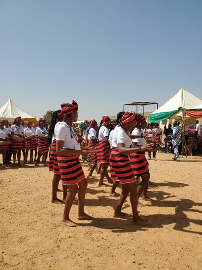 The scene depicts an outdoor event in Bar Gada, Nigeria, characterized by a clear blue sky and light brown dirt ground. Approximately fifteen to twenty individuals, predominantly women, are engaged in a synchronized performance or dance. They wear uniform attire: white short-sleeved tops, short skirts with horizontal red and black stripes, and matching red and black patterned headwraps. Most participants are barefoot. Several individuals in the foreground hold small, dark objects in their hands, consistent with props or instruments. In the background, two white canopy tents are visible; the tent on the right is draped with green and red fabric. A male individual, dressed in blue and white patterned clothing, stands near this tent. Other indistinct figures are present further in the background, observing the activity.
