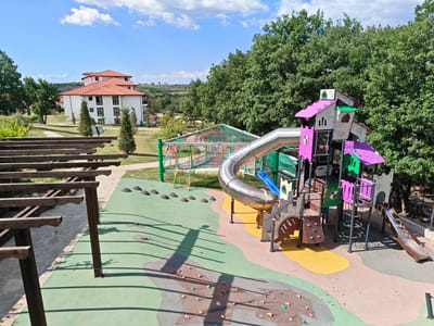 The image depicts a playground in Starosel, Bulgaria, during daylight. The playground features a variety of play equipment, including a large, multi-story structure with a metal slide, climbing walls, and smaller slides. The ground is covered with a mix of green artificial turf and padded surfaces. To the left of the frame, a wooden pergola provides shade. In the background, there is a large building with a red-tiled roof, surrounded by trees and greenery. The sky is blue with scattered clouds. There are no people visible in the immediate scene.