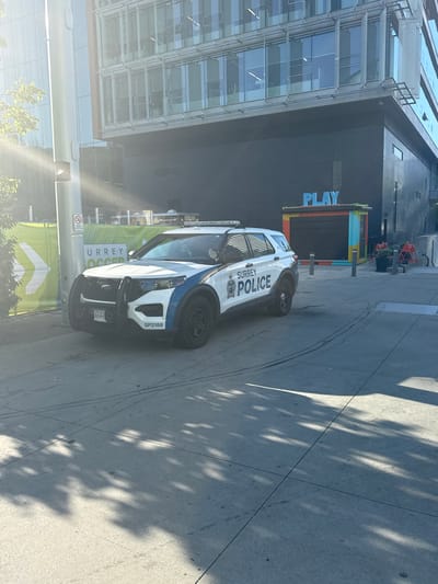 A Surrey Police SUV is parked on a concrete sidewalk in front of a modern building. The building has a dark facade and large windows with a blue "PLAY" sign visible. A green banner with white text and a white arrow is behind the police vehicle. The sun is shining, casting shadows from trees onto the ground. The SUV is white with blue and black markings, displaying "SURREY POLICE" on the side and "SP2188" on the rear bumper. A license plate with "A64-9" is visible on the front of the vehicle. A metal pole with a sign is to the left of the SUV. The environment appears to be an urban setting in Surrey, Canada.