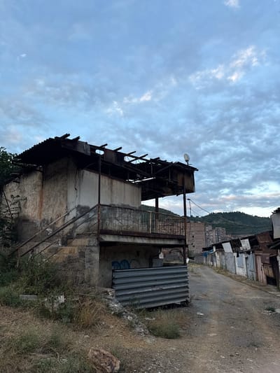 The image depicts a view of a dilapidated building structure with a water tank on top, against a backdrop of a cloudy sky and a hillside with buildings. The structure appears to be made of concrete and metal, with visible signs of damage and decay, including exposed beams, rusted metalwork, and a partially collapsed roof. A satellite dish and a street lamp are mounted on the structure. The surrounding area shows more buildings, some appearing unfinished or in disrepair, and a dirt road. Vegetation, including dry grass and some trees, grows around the base of the structure. The overall scene suggests an urban or semi-urban environment that has seen better days. The time of day appears to be late afternoon or early evening, given the soft light and the color of the sky. The weather seems overcast, with scattered clouds. The location is identified as Kapan, Armenia. There is no visible text in the image.