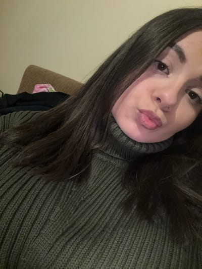 A close-up, selfie-style image captures a young female indoors in Radomir, Bulgaria. The subject has long, dark brown hair and dark eyes, and is wearing an olive-green ribbed turtleneck sweater. Her head is tilted slightly to her left, and her lips are puckered in a "kiss" expression, directed towards the camera. The background reveals a light-colored wall and the upper portion of a tan, upholstered furniture item, possibly a couch or headboard, upon which dark fabric and a pink patterned item are visible.