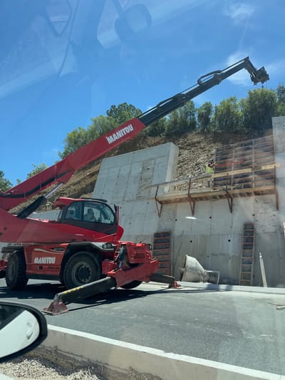 The image shows a construction site with a large red Manitou telehandler in the foreground. The telehandler's boom is extended upwards and to the right, with its attachment obscured. The machine has "MANITOU" and "MRT 2550+" branding visible. Inside the cabin, two individuals are seated. A concrete retaining wall is under construction behind the telehandler, with wooden formwork attached to its surface. A person wearing a yellow vest and hard hat is working on the formwork. The ground is a mixture of asphalt and gravel. In the background, a wooded hillside rises under a clear blue sky. A concrete form or mold lies on the ground in front of the wall. The scene appears to be located in Lastva Grbaljska, Montenegro.