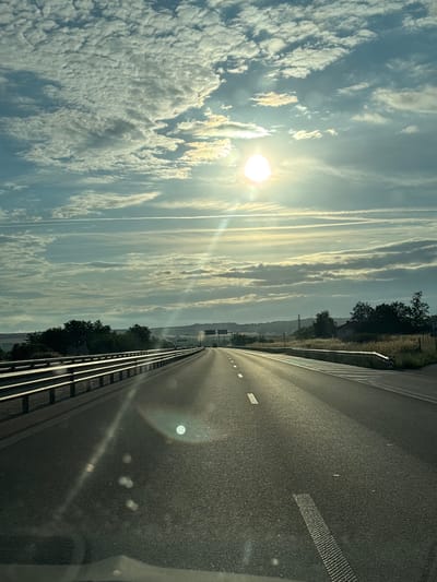 The image shows a highway with a metal guardrail on the left and dry grass and trees on the right. The sun is low in the sky, casting a bright glare and creating lens flare effects on the road. The sky is filled with scattered clouds, some appearing wispy and others more textured. The road markings, including dashed white lines and reflective studs, are visible. In the distance, there are more trees and a gentle slope of land. The scene is captured from a perspective that suggests movement, likely from inside a vehicle traveling on the highway. The environment appears to be rural or suburban.