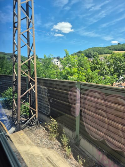 The image depicts a view from a train traveling through the city of Kobern-Gondorf, Germany. A tall, rusted metal pylon is visible on the left side of the frame, positioned close to the train. A long, horizontal fence, likely a sound barrier, runs along the right side of the track. Beyond the fence, a hillside covered in lush green trees and vineyards is visible. Several buildings, typical of a German town, are scattered among the trees on the hillside. The sky is a bright blue with scattered white clouds, suggesting fair weather. The overall scene conveys a sense of travel through a scenic, rural area of Germany.