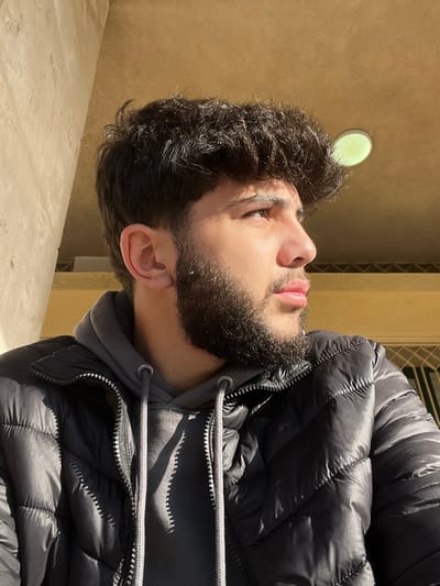 A young man with dark, curly hair and a trimmed beard looks upwards. He wears a grey hooded sweatshirt beneath a black puffer jacket. The background features a beige wall with a circular light fixture and a metal grate. Sunlight highlights his face and casts shadows on the jacket. The image is rotated counterclockwise.