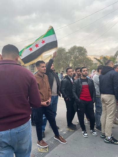 The image displays an outdoor gathering of approximately 15 to 20 individuals, primarily young men, situated on a paved street in Al-Qanawat Municipality, Syria. The scene is illuminated by an overcast sky. In the mid-ground, a man wearing a dark jacket holds a flag horizontally; the flag consists of three horizontal stripes (green, white, black from top to bottom) and three red five-pointed stars centered on the white stripe, bordered by a gold fringe. In the left foreground, a man is seen from the back, wearing a maroon sweater and blue jeans. To his right, a man in a brown collared shirt and dark cargo pants stands on a yellow tactile paving block while holding a smartphone. Other individuals in the mid-ground and background are dressed in various casual attire, including dark jackets, sweaters, and jeans. The ground is paved, with additional yellow tactile paving blocks visible. Overhead, multiple power lines crisscross the sky. The background features light-colored urban buildings, deciduous trees, and a palm tree, with a distant elevated area or hill partially visible. The individuals are standing and appear to be observing the scene, with no discernible active interactions or specific actions beyond their assembly.