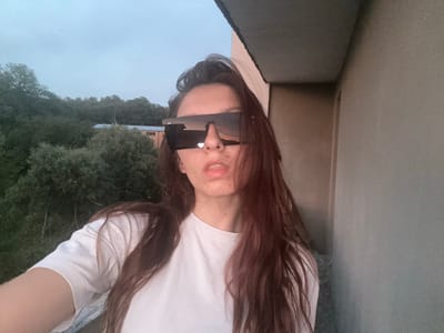 A young woman stands on a balcony, facing the camera. She is wearing a white t-shirt and large, angular sunglasses. Her long, dark hair is loose around her shoulders. Behind her, the sky is overcast, and a dense line of trees is visible in the distance. To the right, a portion of a building wall is visible. The image appears to have been taken in the city of Tryavna, Bulgaria.