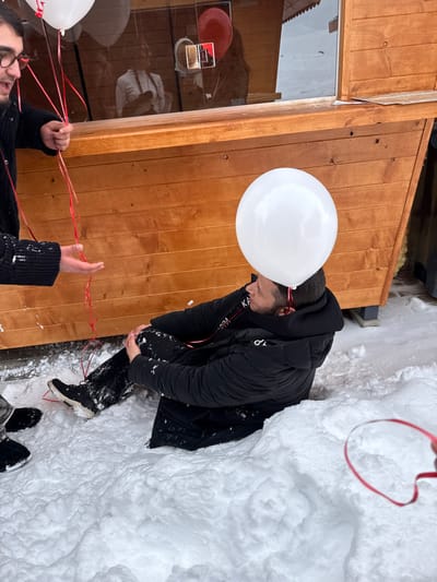 The scene is set in an outdoor, snow-covered environment in Yeghipatrush, Armenia. A male individual is seated directly in deep snow in the center-right of the frame, wearing a black puffer jacket, black pants, and black athletic shoes. Snow adheres to his clothing and footwear. A white balloon rests on his head, with a red ribbon extending from it, partially visible near his neck. He is positioned in profile, looking towards the left. To the left, a second male individual, partially visible, is standing. He wears dark clothing and glasses, holding multiple white balloons with red ribbons in his right hand. His left hand is extended downwards with an open palm. In the background, a light brown wooden structure, possibly a kiosk or small building, features a window. The reflection of an individual, seemingly with braided hair, and a red balloon are visible within the window pane. The ground throughout the visible area is uniformly covered in thick white snow.